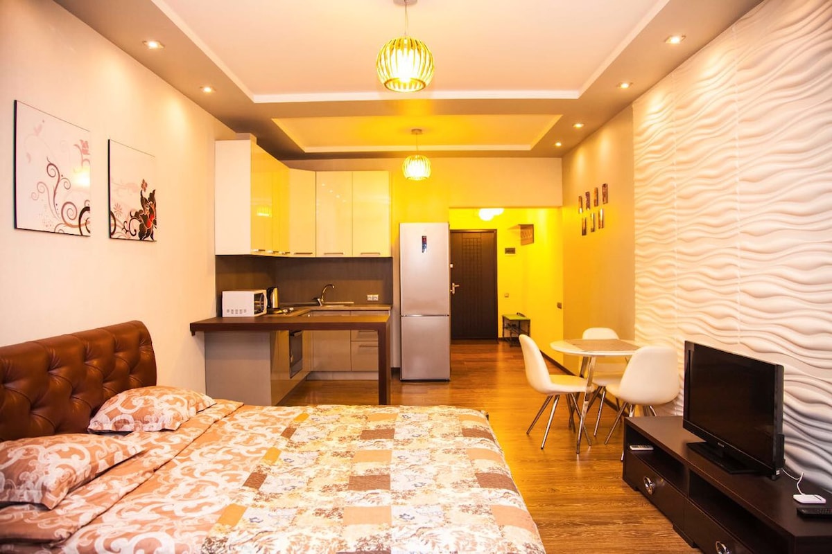 A cozy studio apartment features a queen-sized bed with a decorative floral pattern. A compact kitchen area is equipped with modern appliances, including a refrigerator and microwave. A dining table with white chairs complements the designed walls, while soft lighting creates a relaxing ambiance.