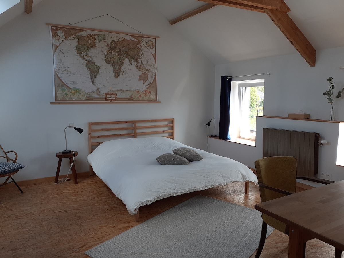 A spacious bedroom features a large bed with white bedding and decorative pillows. A world map is displayed on the wall, while natural light filters through a window adorned with simple curtains. A small table and chairs complement the cozy atmosphere.