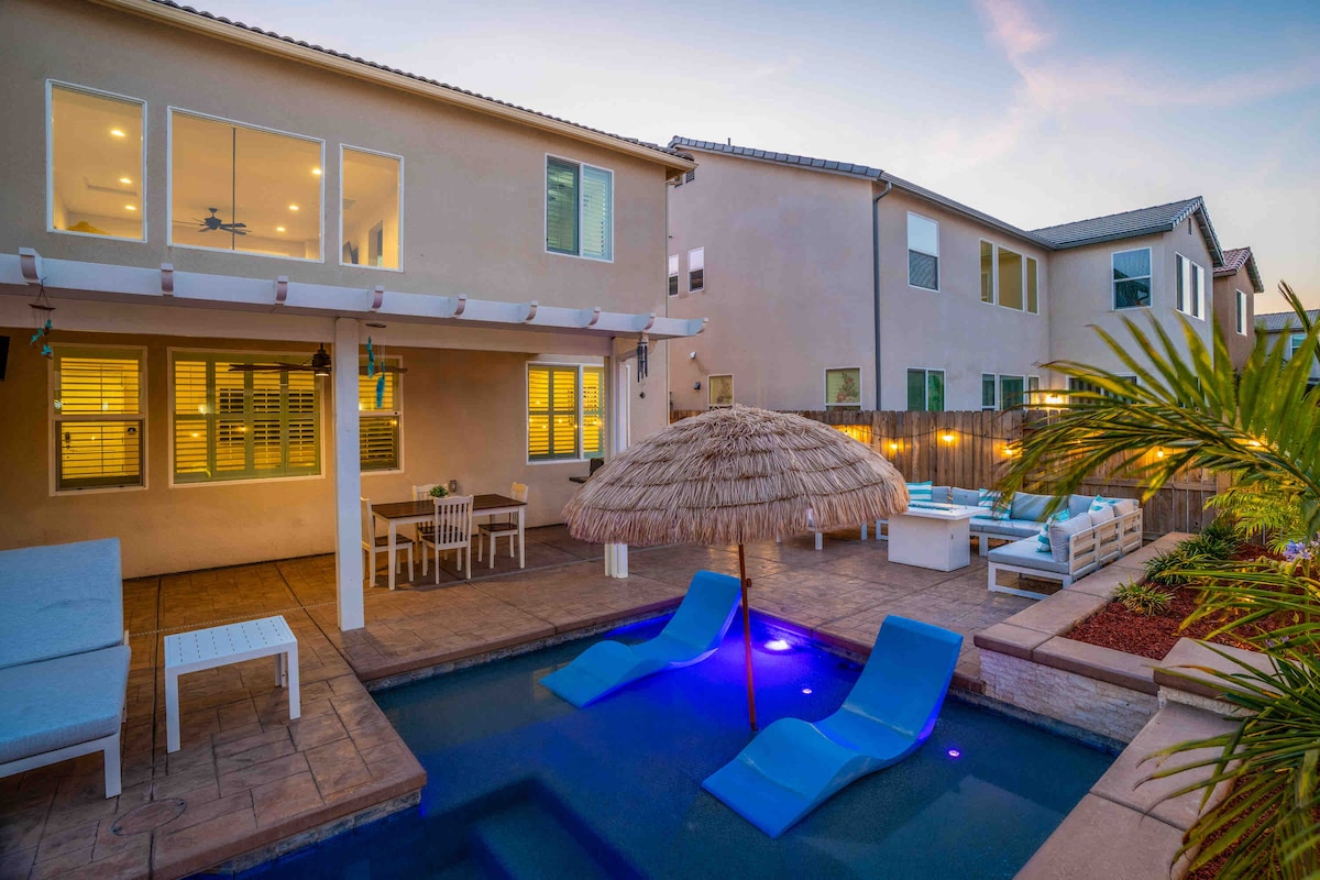 An outdoor area is shown with a heated pool featuring lounge chairs and a thatched umbrella providing shade. The patio includes a dining table and chairs, with ambient lighting enhancing the space. The backdrop features a two-story building with large windows.