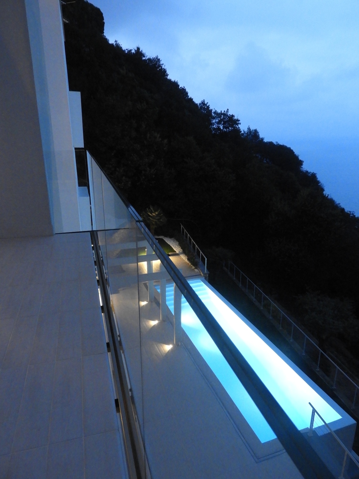 An elevated view of the shared pool illuminated softly at dusk is captured from a balcony. The pool area is framed by landscaped surroundings, with modern railings providing safety and transparency. The ambiance is serene, showcasing a seamless connection to nature.