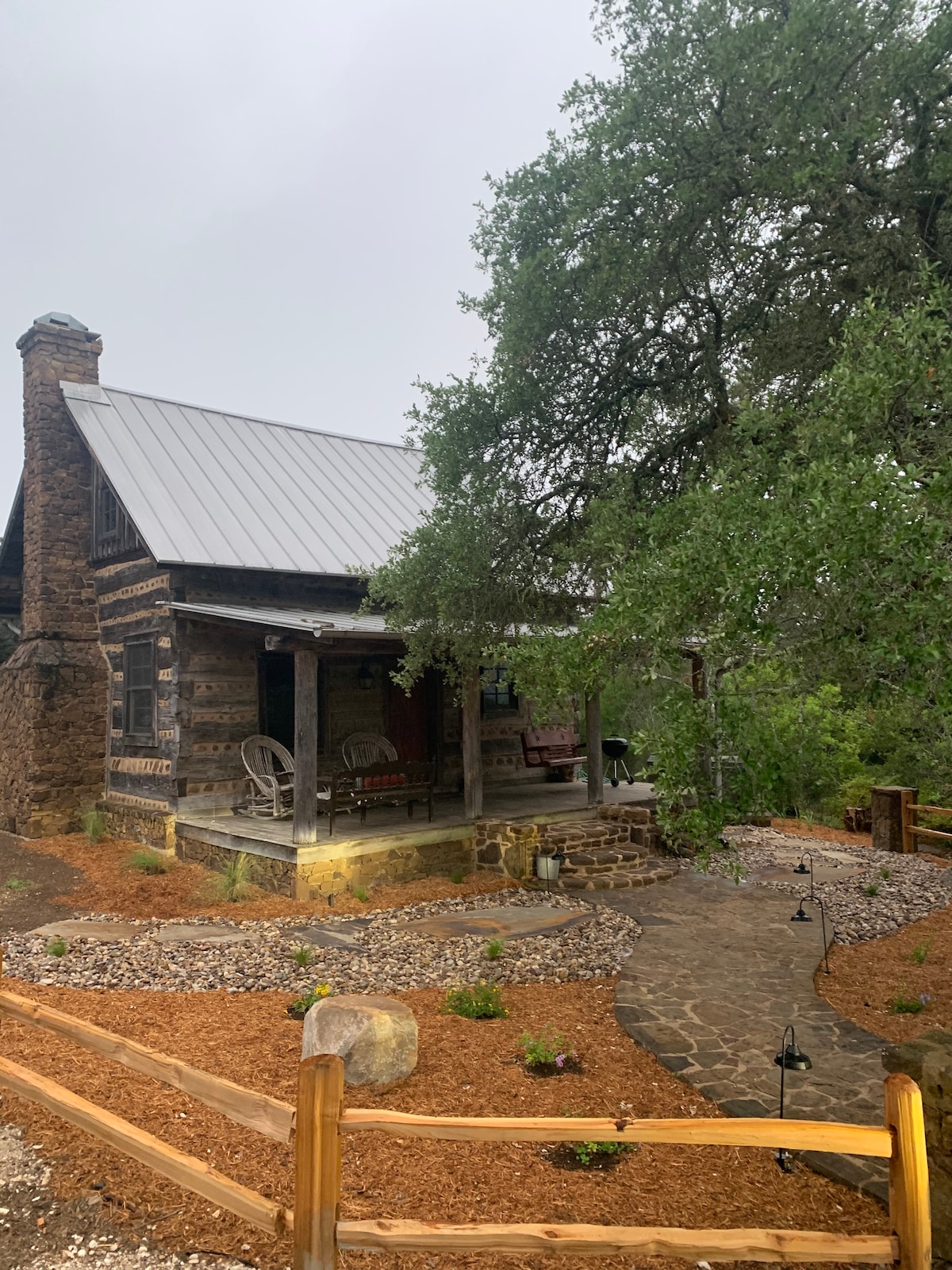 The cabin exterior is framed by a natural landscape, showcasing the authentic log construction and a sloped metal roof. A welcoming porch features two rocking chairs, while a flagstone pathway leads through a well-maintained garden area, bordered by wooden fencing and decorative lights.