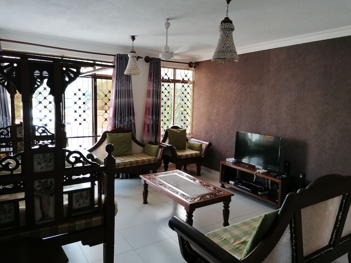 A spacious living area features traditional wooden furniture with patterned cushions. Two intricately designed chairs and a matching sofa surround a wooden coffee table. Natural light filters through large windows adorned with patterned curtains, while a flat-screen TV is placed on a media unit against the wall.