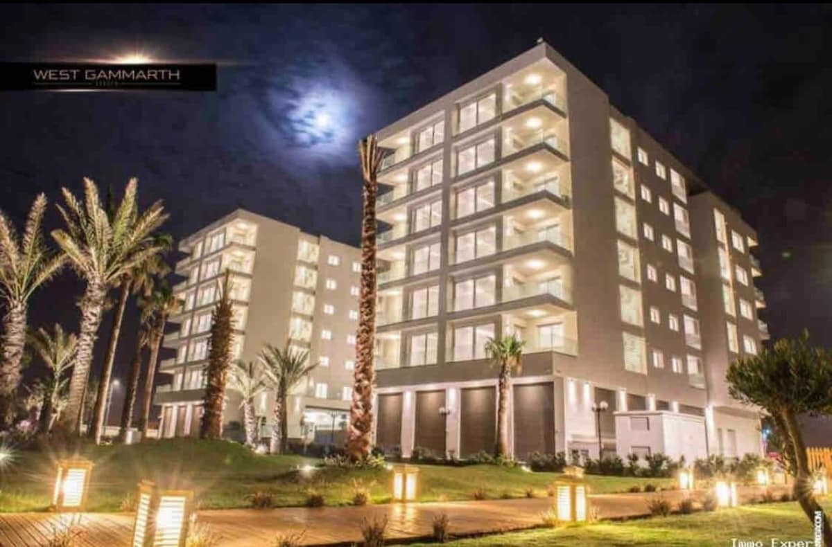 Two modern buildings are illuminated against the night sky, showcasing multiple balconies and large windows. Palm trees are positioned in the foreground, and soft lighting highlights the landscaping, creating a serene ambiance.