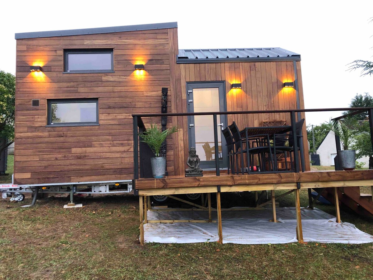 A wooden exterior of a tiny house is highlighted by warm outdoor lighting. A small deck features a dining table with chairs and potted plants. Large windows provide natural light, while the modern design complements the tranquil setting.