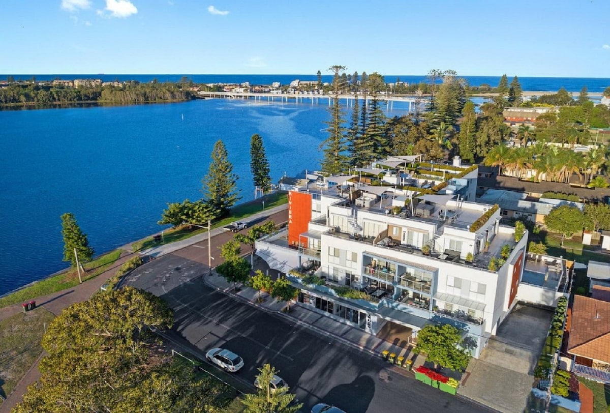 An aerial view of the modern apartment complex situated by the water's edge, featuring a rooftop terrace. The surrounding landscape includes trees and a clear blue sky, with the shimmering lake and distant shoreline visible in the background.