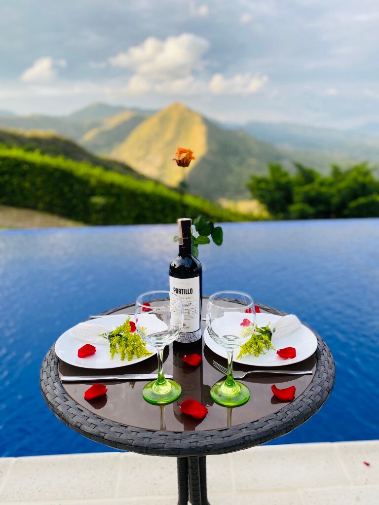 A table is elegantly set with two wine glasses and plates, placed beside a bottle of wine. Red rose petals are scattered across the table, overlooking a serene infinity pool and lush mountains in the background, emphasizing a romantic outdoor dining experience.