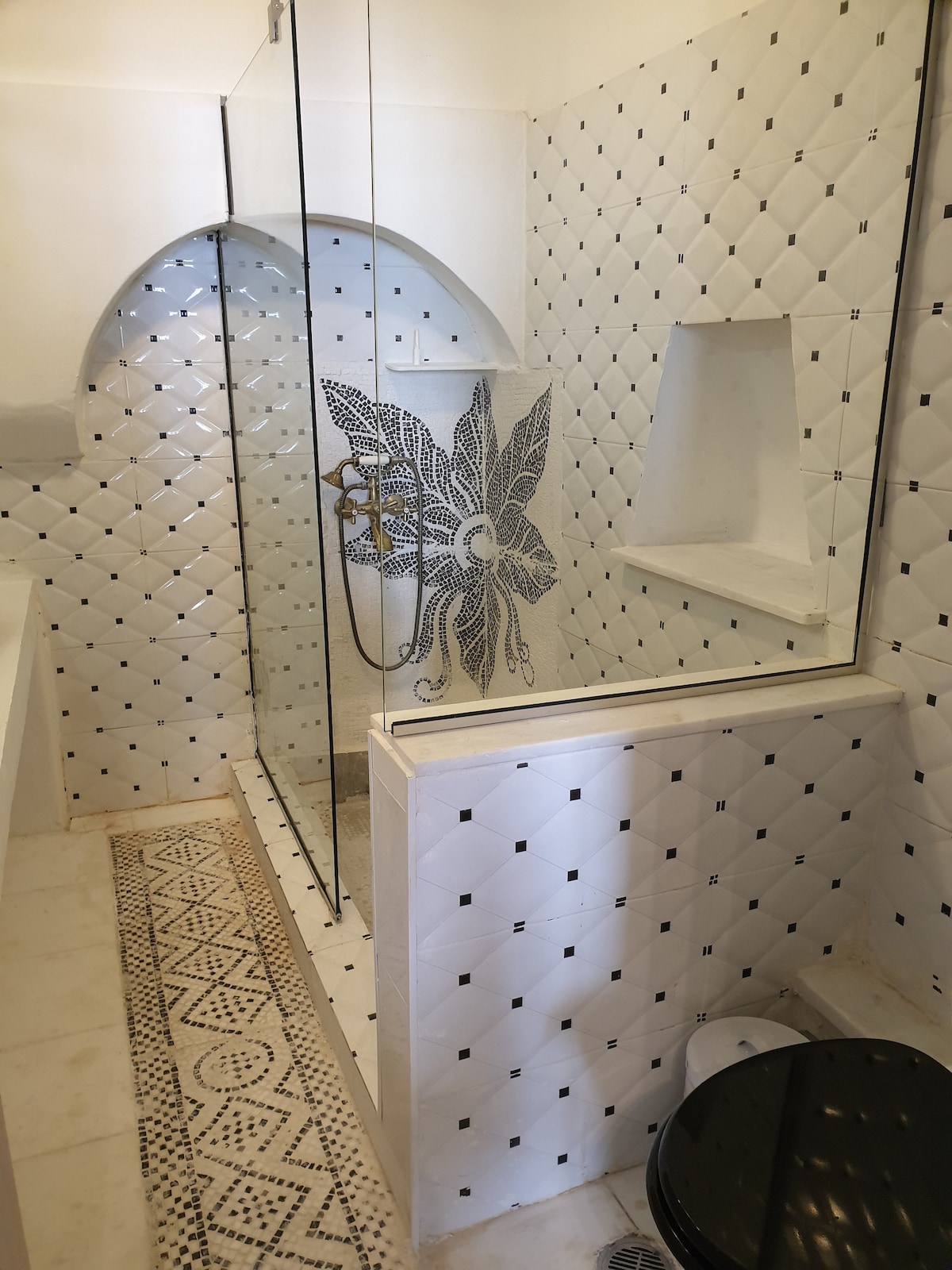 A modern bathroom features a glass shower with intricate tile work, including a decorative design and an arched nook. A minimalist black toilet complements the white interior, which is highlighted by patterned flooring and glossy wall tiles in a diamond layout.