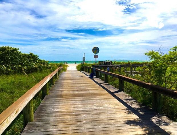 A wooden boardwalk extends through lush greenery, leading towards a sandy beach and the Gulf of Mexico. A clear blue sky is visible above, with a few clouds scattered. Signs are positioned along the pathway, guiding visitors to the beach area.