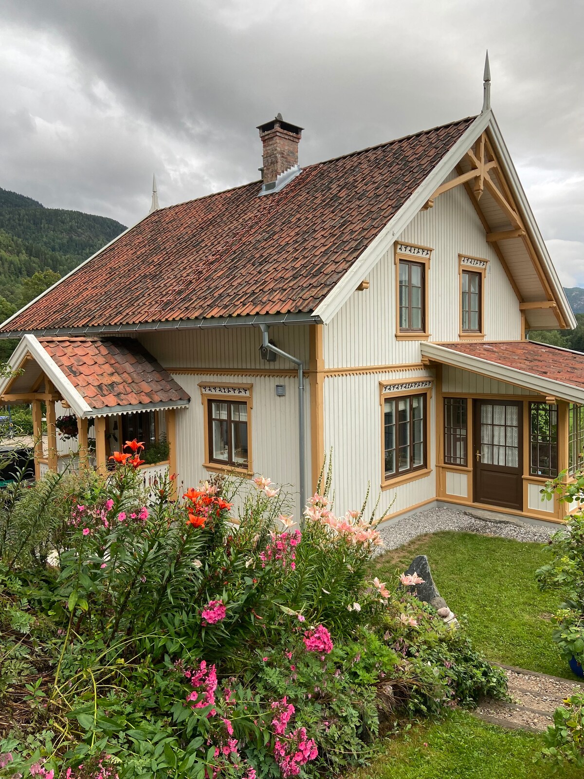 A charming house is depicted with a distinctive roof and decorative wooden details. Colorful flowers bloom in the foreground, providing a natural contrast to the light-colored siding. Surrounded by lush greenery, the structure is set against a backdrop of gently rolling hills.