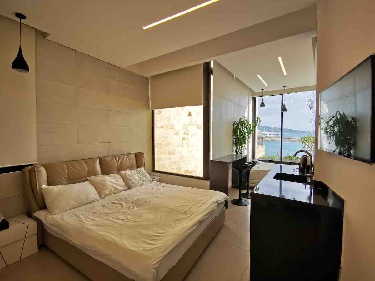 A spacious room features a large bed with neutral-toned bedding. Natural light fills the area through large windows, providing a view of the Mediterranean. A modern desk and chair are positioned by a window, while a sleek black counter adds functionality.