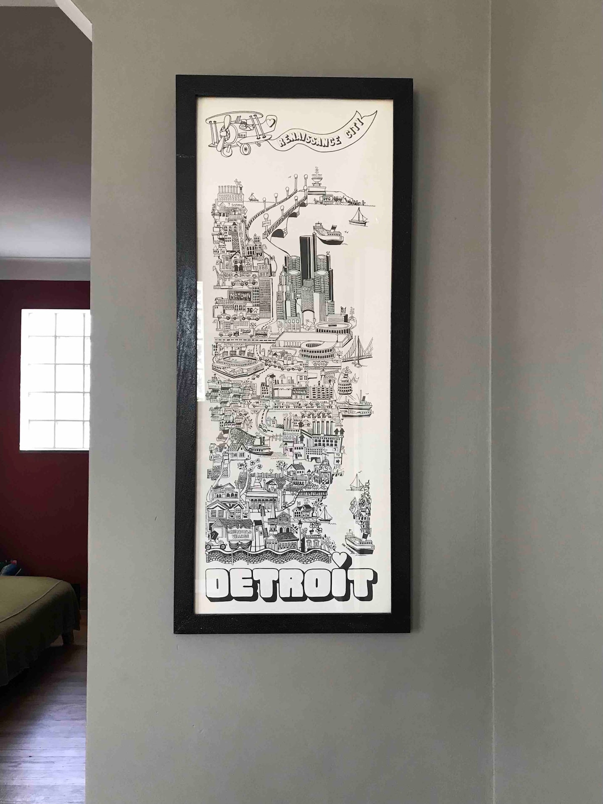 A framed illustration showcasing a detailed map of Detroit is mounted on the wall. The artwork features various city landmarks and highlights, with the word 'DETROIT' prominently displayed at the bottom. A soft light from a nearby window softly illuminates the area.