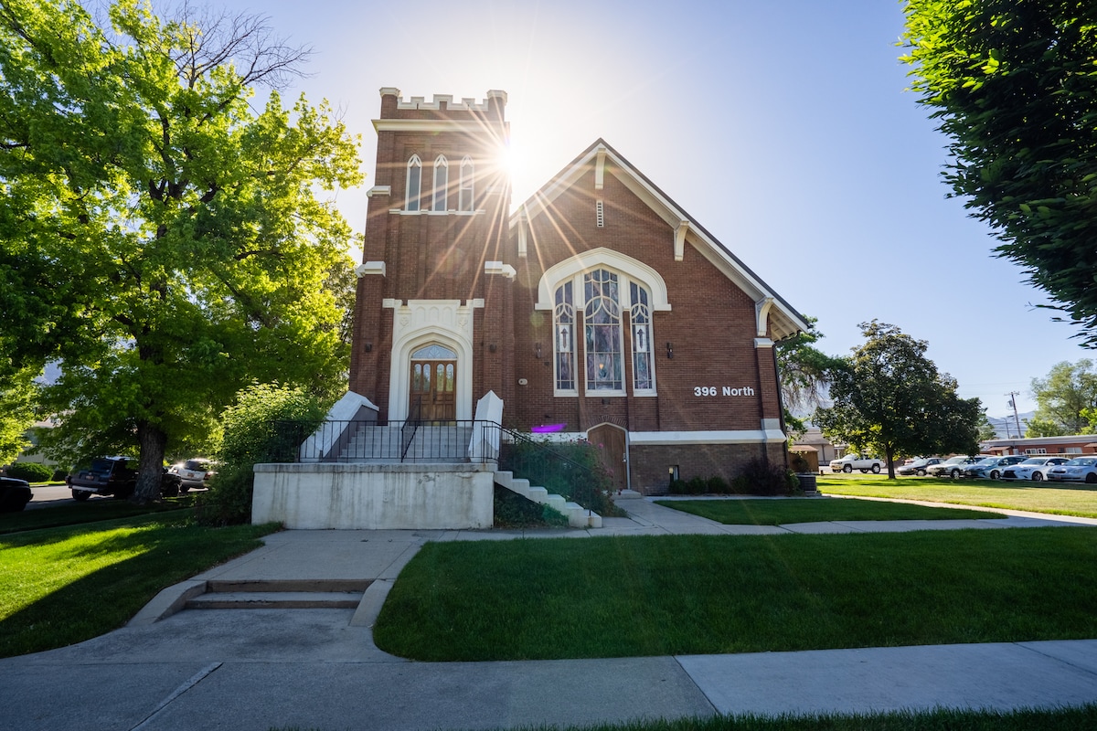 The exterior of a stately red brick building is displayed, featuring tall, elegant stained glass windows. Sunlight streams from behind the structure, casting a warm glow on the well-maintained lawn and pathways. Lush green trees frame the scene, enhancing the surroundings.