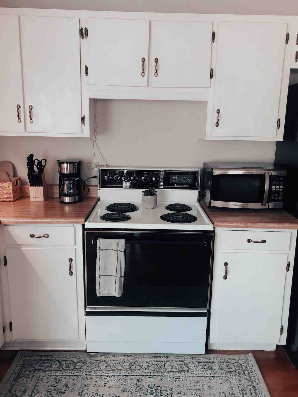 A functional kitchen area includes a black stove with four burners, flanked by a coffee maker and a microwave. White cabinetry offers ample storage, while a wooden countertop provides cooking space. A rug is positioned beneath, lending a touch of warmth to the setting.