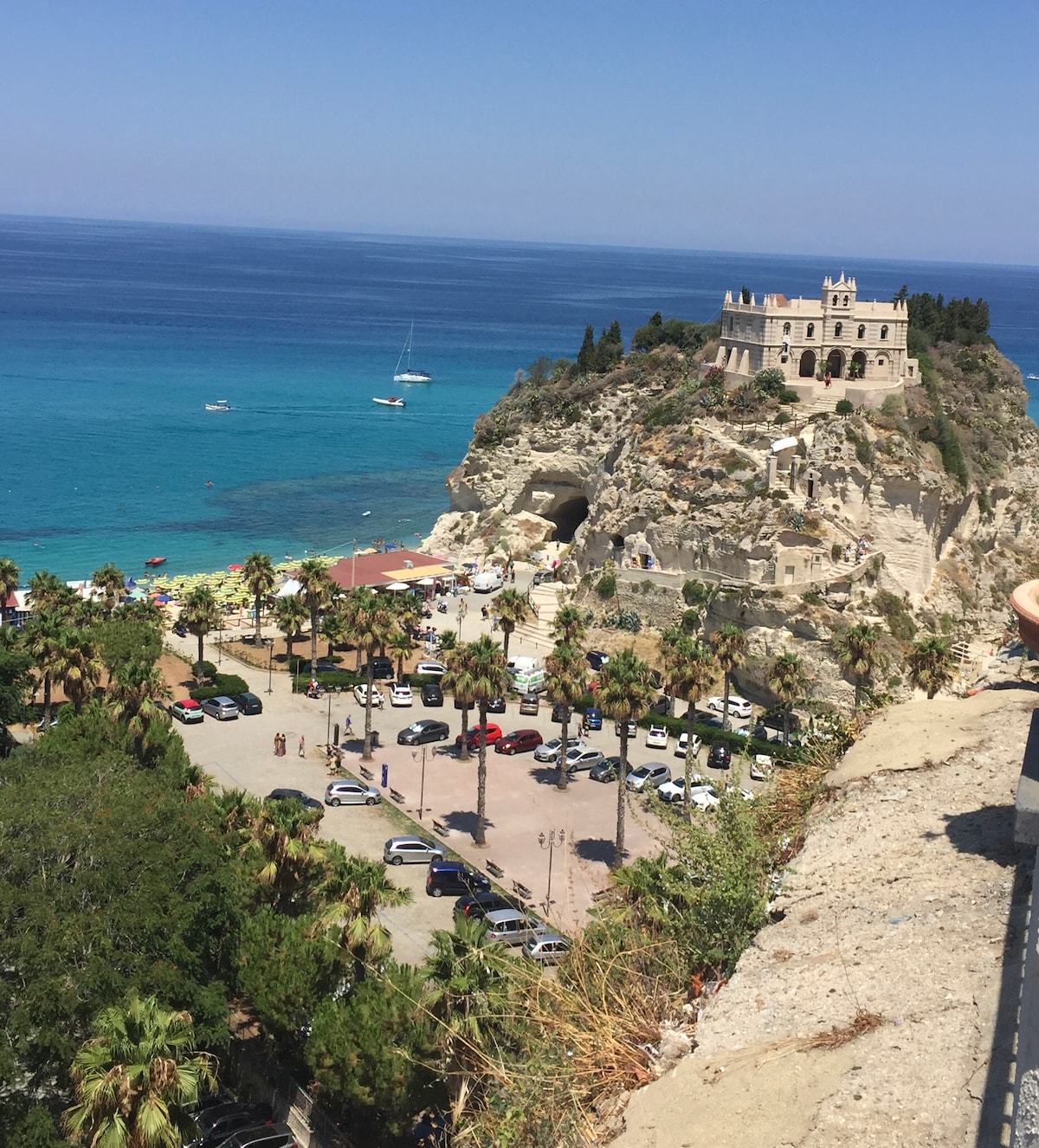 A panoramic view of a rocky coastline features a historic building perched atop a cliff overlooking the sea. The turquoise water and sandy beach are visible below, along with scattered boats and a line of palm trees. An area of parked cars is seen in the foreground.