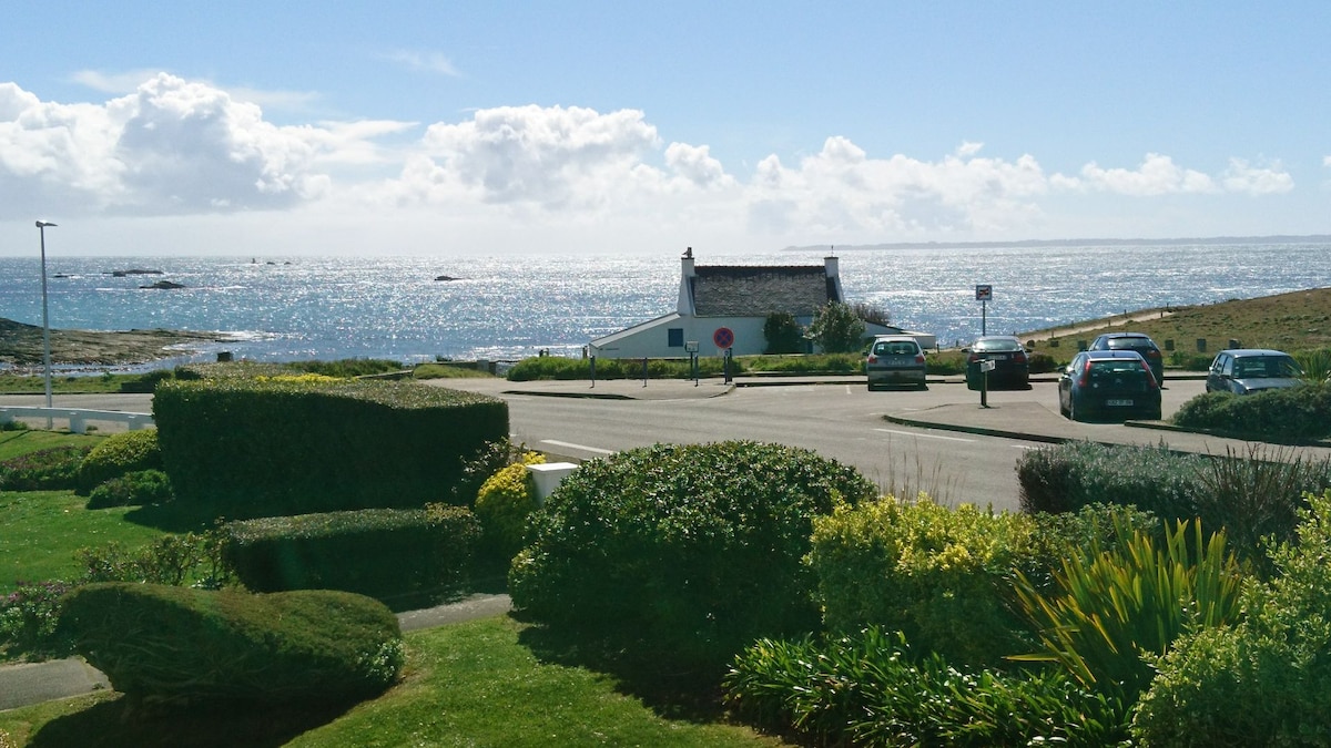 The image captures a view of the sea, showcasing shimmering waters under a clear sky. Lush green shrubs are visible in the foreground, and a white building can be seen in the distance. Several parked cars line the road, contributing to the serene setting.