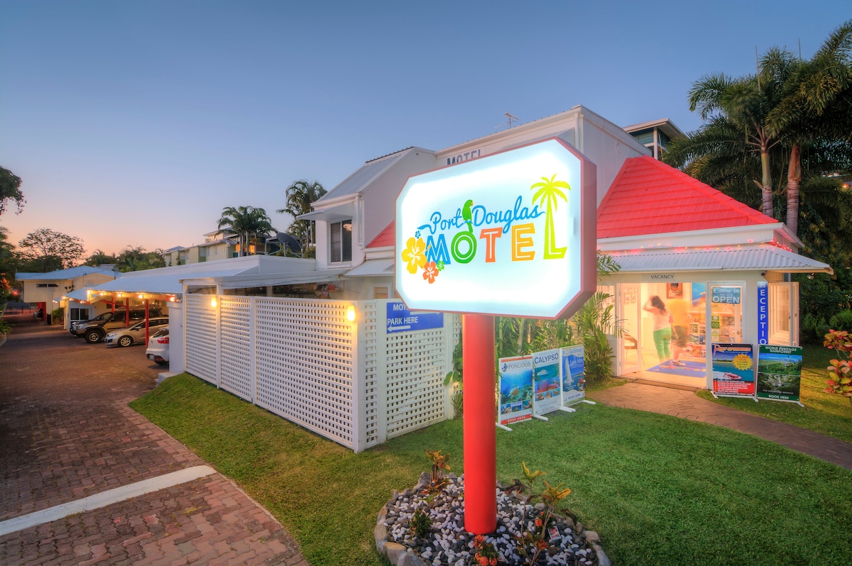 The exterior of the Port Douglas Motel is showcased, featuring a colorful, illuminated sign with a tropical theme. Lush greenery surrounds the property, complementing the welcoming atmosphere. The well-maintained façade is highlighted against a sunset sky, providing a glimpse of the nearby parking area.