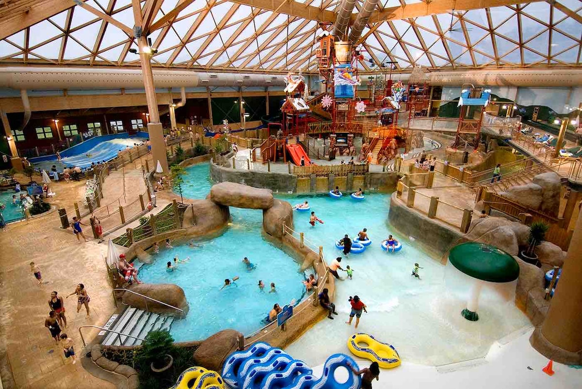 An expansive indoor water park features several pools, water slides, and play areas. Guests of various ages can be seen enjoying the attractions, while clear roofs provide natural light to the vibrant space. Seating areas are placed around the water features for relaxation.