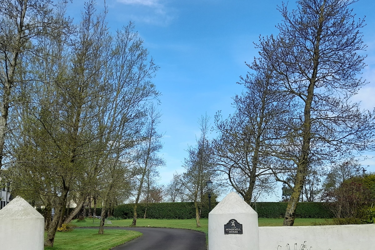 The entrance to the property is framed by neatly manicured lawns and trees. A clear blue sky can be seen overhead, and a stone marker is visible at the gate, indicating the property's name. The roadway leads into a tranquil rural setting.
