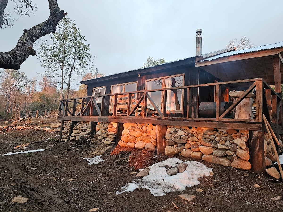 The exterior of a rustic cabin is shown, featuring a wooden deck supported by large stones. Soft warm light emanates from the windows, highlighting the inviting atmosphere. Surrounding vegetation and remnants of snow can be seen, indicating a serene natural setting.