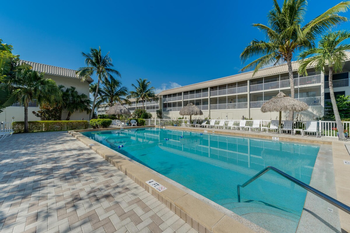 A clear view of a swimming pool is presented, lined with lounge chairs and shaded by palm trees. Umbrellas provide additional shade beside the pool, and the surrounding area features well-maintained landscaping. The sun shines brightly against a clear blue sky.
