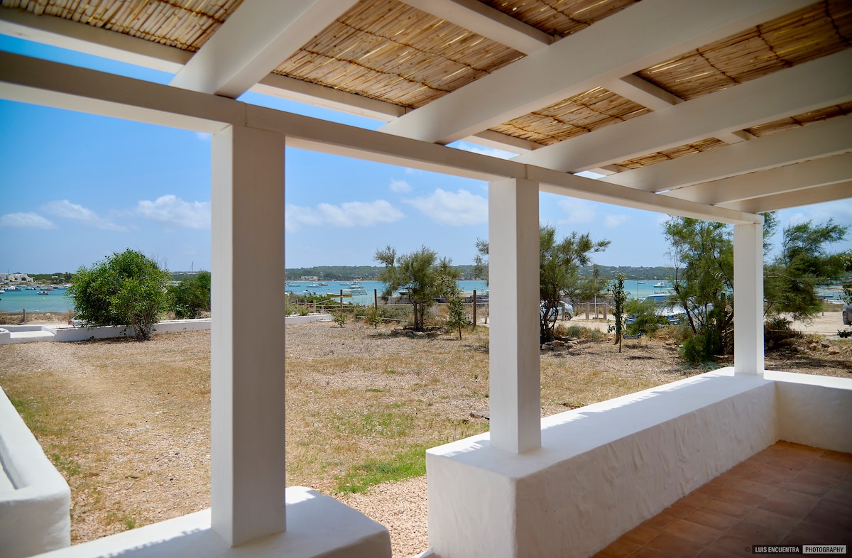 A spacious terrace is visible, featuring a thatched roof supported by white pillars. The serene view includes grassy land leading to a waterfront scene, where calm waters shimmer under a clear blue sky.