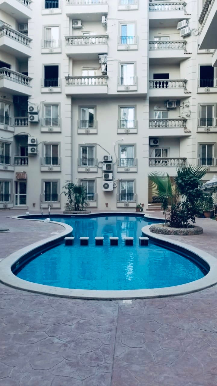 A serene outdoor swimming pool is centrally located within the building's courtyard, surrounded by well-maintained greenery. The pool features a curved design, with sun loungers positioned nearby for relaxation. The surrounding architecture includes multiple balconies and air conditioning units.