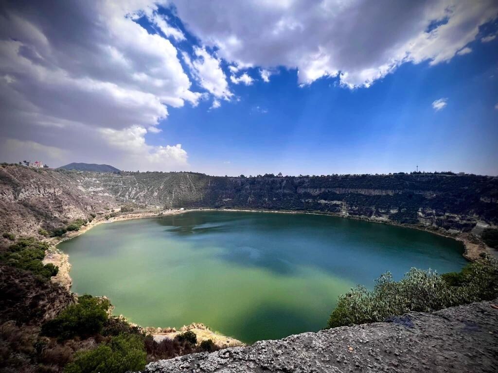 A tranquil lagoon is depicted, surrounded by rugged cliffs and lush greenery. The water reflects a vibrant green hue under a partly cloudy sky, creating a serene atmosphere. The depth of the lagoon is accentuated by the surrounding terrain, inviting exploration of the natural surroundings.
