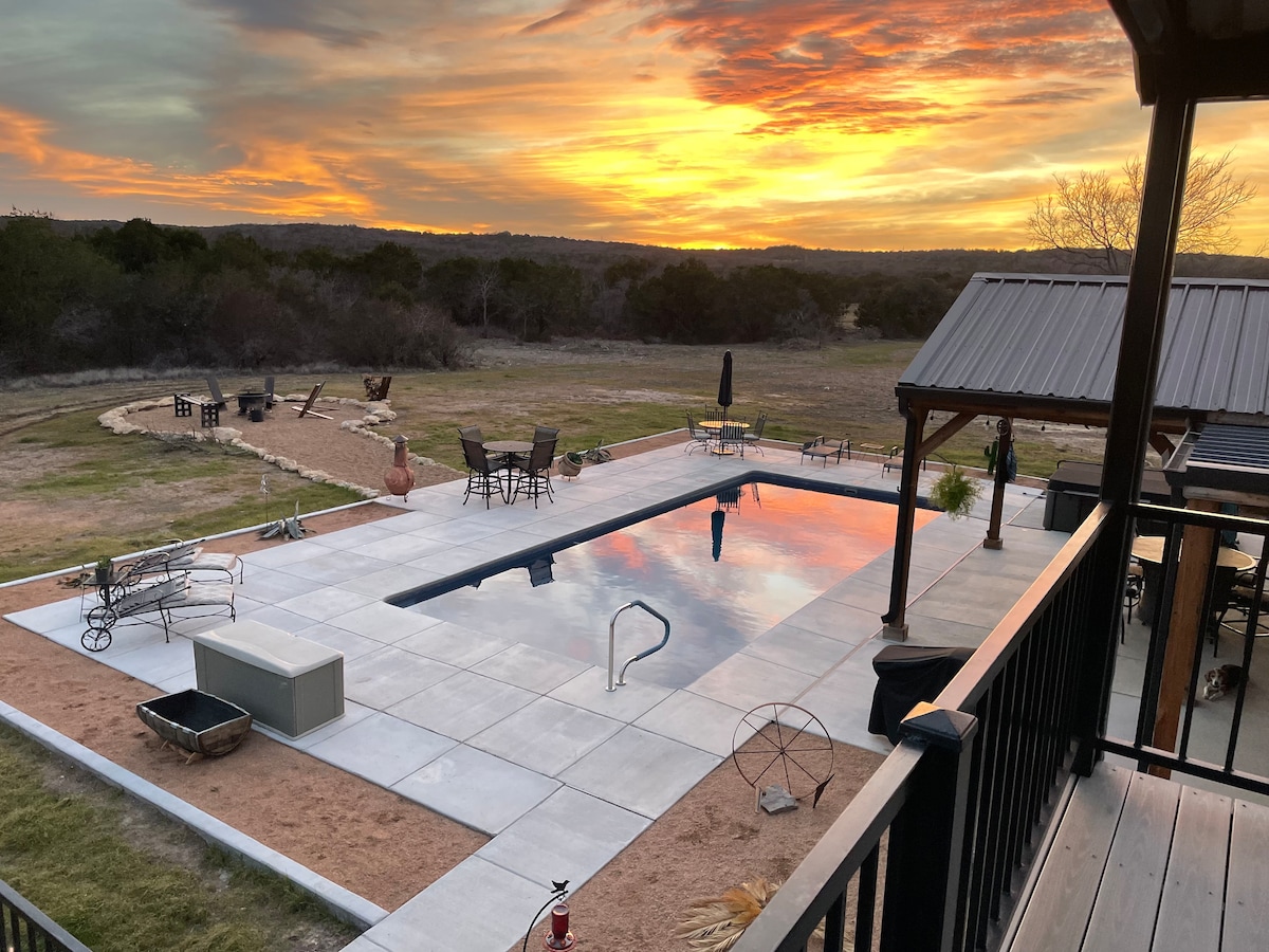 A spacious patio encompasses a large swimming pool reflecting the sunset, offering a tranquil setting. Lounge chairs and outdoor furniture are positioned around the pool, with views of the surrounding landscape and a path leading to additional seating areas.