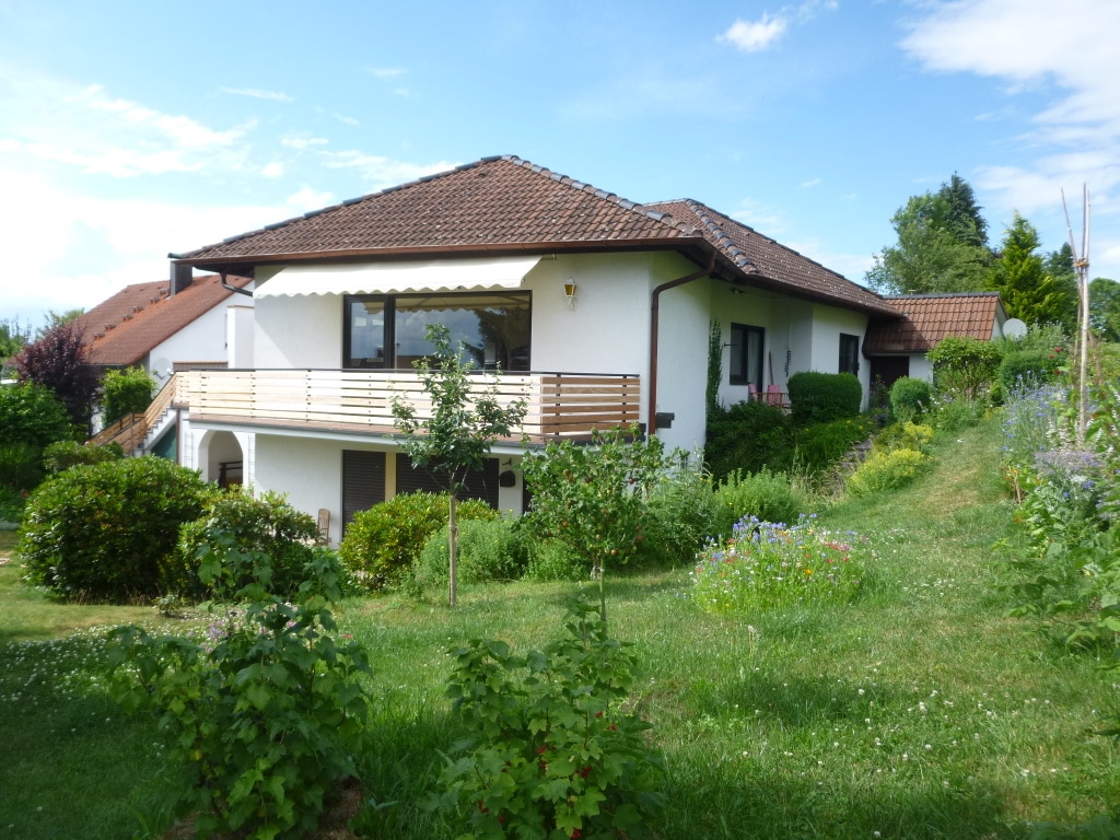 The exterior view of a single-story house is presented, surrounded by lush greenery and colorful flowering plants. A small balcony extends from the main floor, and well-maintained shrubs frame the entrance. The soft slope of the garden adds depth to the landscape.