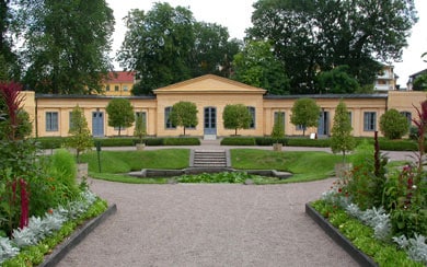 An elegant building with a symmetrical facade is framed by well-maintained gardens. Low hedges and ornamental trees create a harmonious layout. Pathways of light gravel lead towards the entrance, while flower beds featuring a variety of plants add color and texture to the surrounding landscape.
