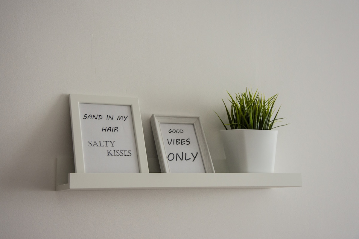 A minimalist shelf displays two framed prints and a potted plant. The prints feature simple text in bold fonts. One print includes the phrase 'Sand in My Hair, Salty Kisses,' while the other reads 'Good Vibes Only.' The potted plant adds a touch of greenery.