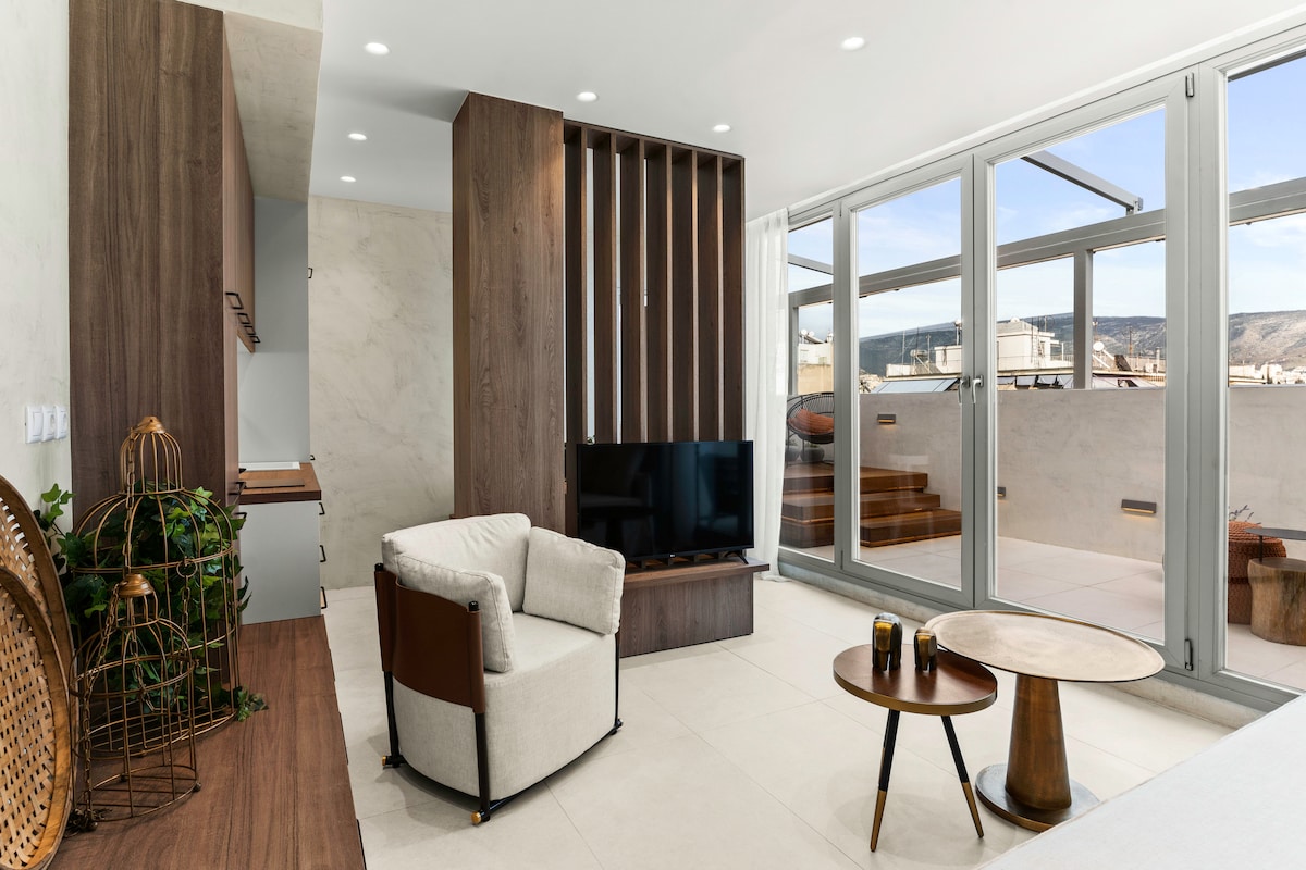 A modern living space is showcased, featuring a light-colored armchair next to a small round table. A flat-screen TV is mounted on a wooden panel. Large glass doors open to a scenic view, allowing natural light to illuminate the room.