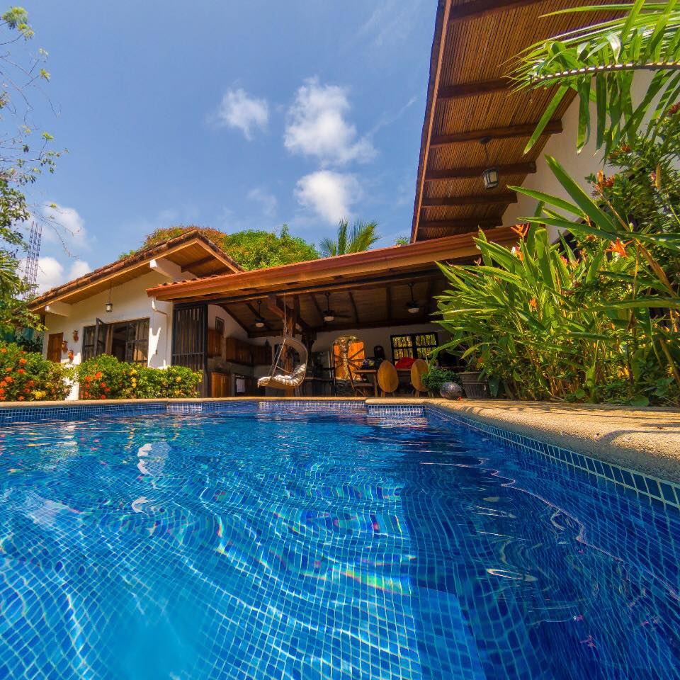 The image captures a sparkling blue pool, reflecting the clear sky and surrounded by vibrant tropical greenery. A charming villa structure is visible in the background, featuring a thatched roof and open spaces, inviting a sense of relaxation and connection with nature.