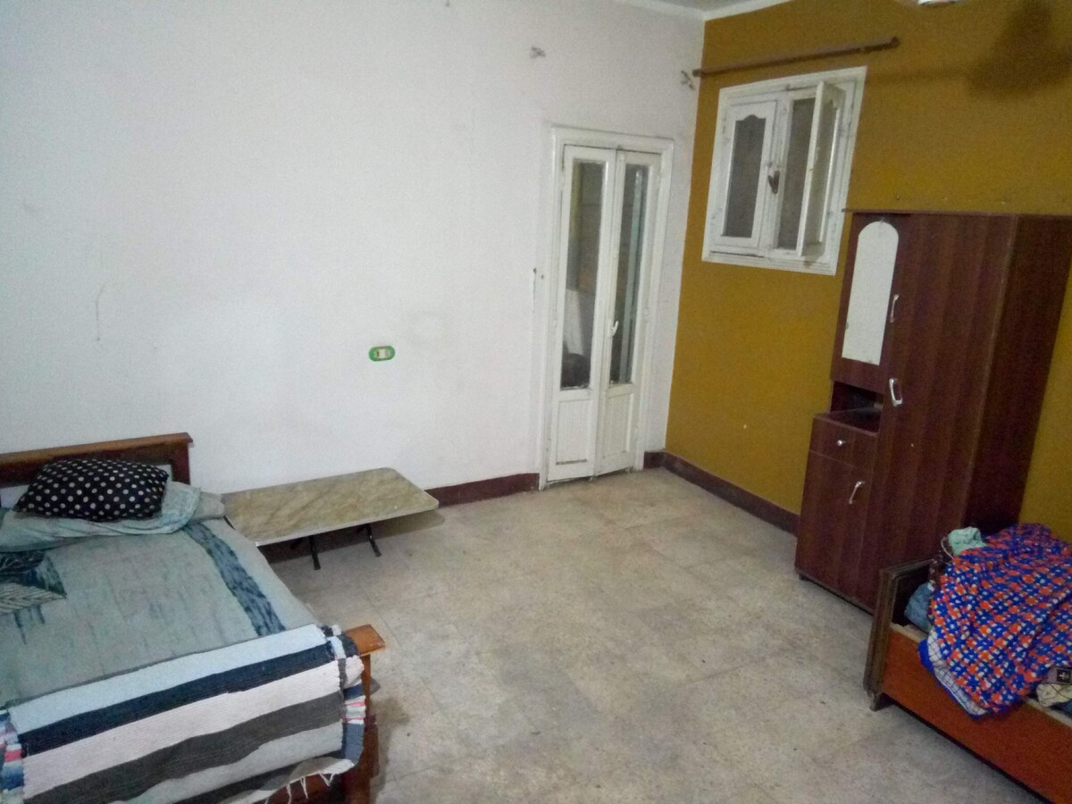 A room featuring two beds, one with a patterned blanket. A small folding table is positioned nearby. The walls are painted in light colors, with one wall highlighted in a warm brown tone. A window allows natural light, complementing the space.