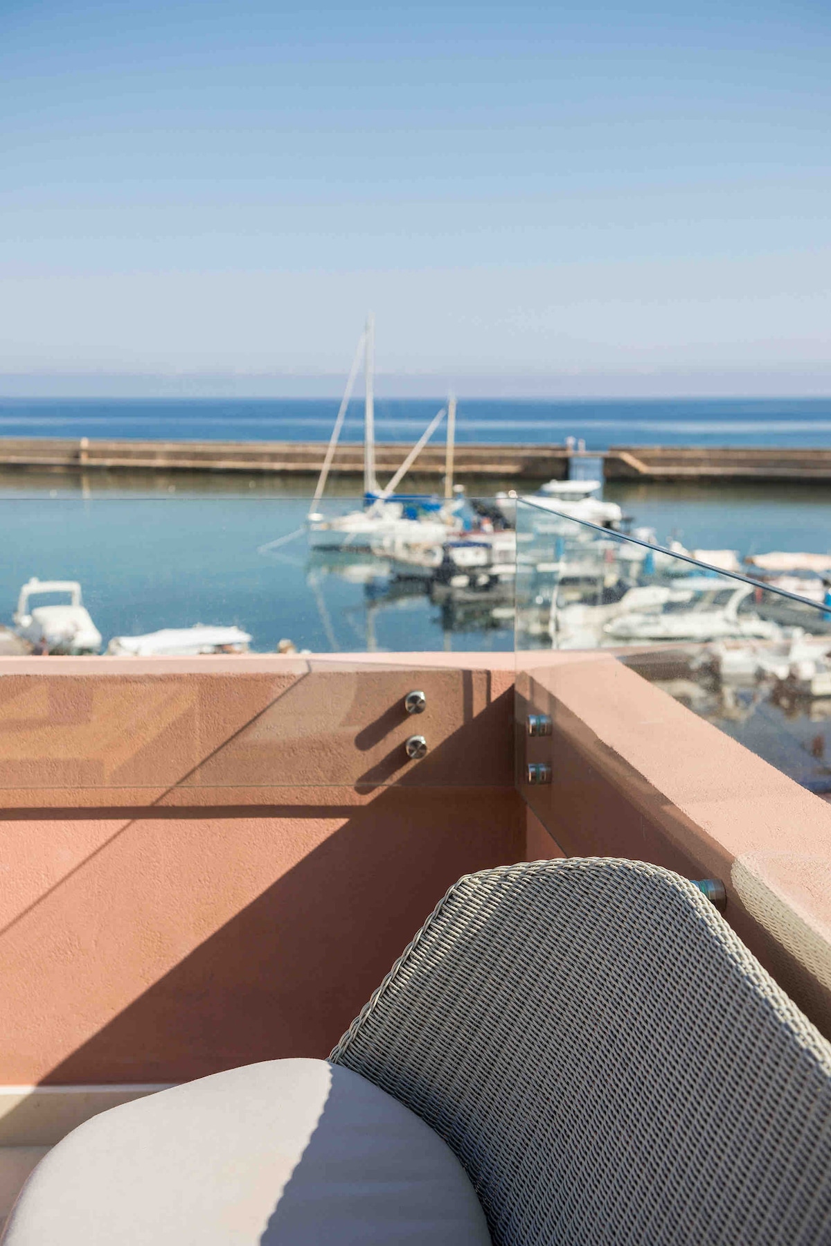 A section of the furnished terrace is visible, featuring a comfortable chair. Beyond, the vibrant blue waters of the Venetian Harbour are seen, with several boats anchored. The shoreline and distant horizon create a serene backdrop under a clear sky.