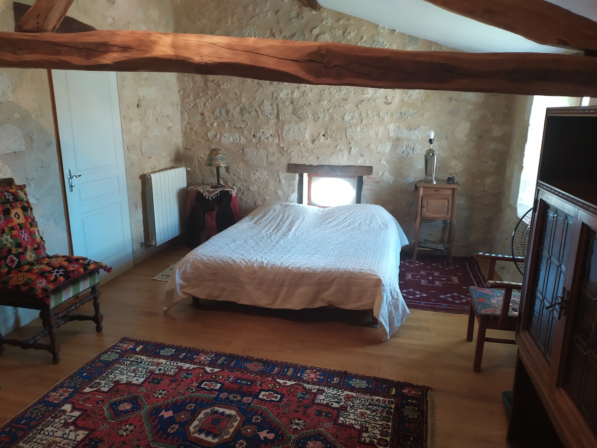 A bright bedroom features exposed stone walls, wooden beams, and a spacious bed covered with a white blanket. Traditional rugs add warmth to the wooden floor. A small wooden nightstand stands beside the bed, and a decorative chair complements the inviting space.