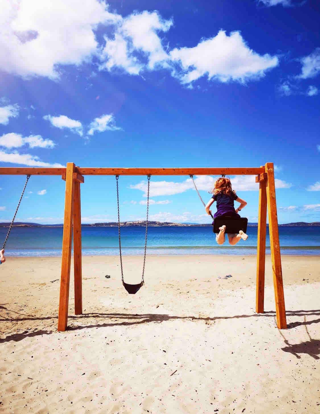 A wooden swing set is positioned on a sandy beach, framed by expansive blue skies and gentle clouds. The ocean stretches in the background, with soft waves lapping at the shore, creating a serene atmosphere. One child is captured mid-swing, adding a dynamic element to the scene.