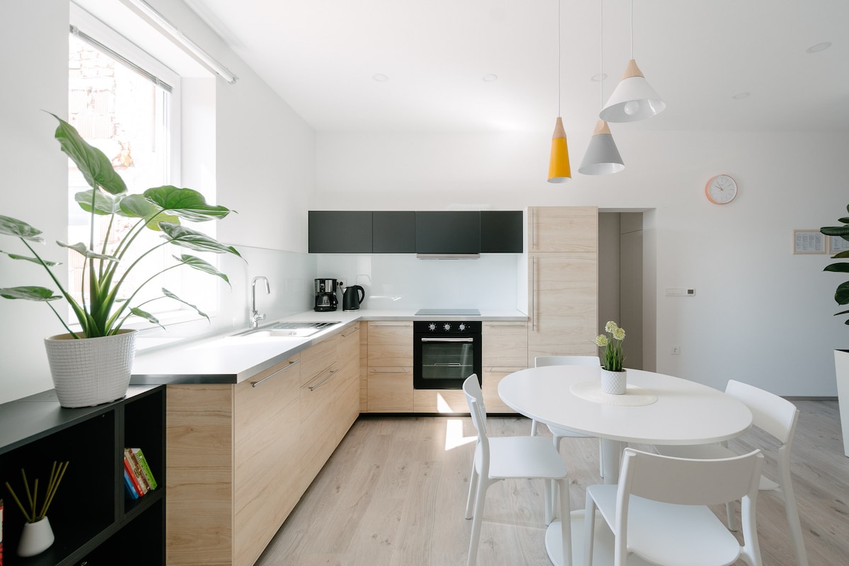 A bright kitchen space features modern cabinetry in light wood and black finishes. A dining area with a round table and white chairs is accompanied by a potted plant. Kitchen appliances, including an oven and coffee machine, are neatly arranged along the counter.