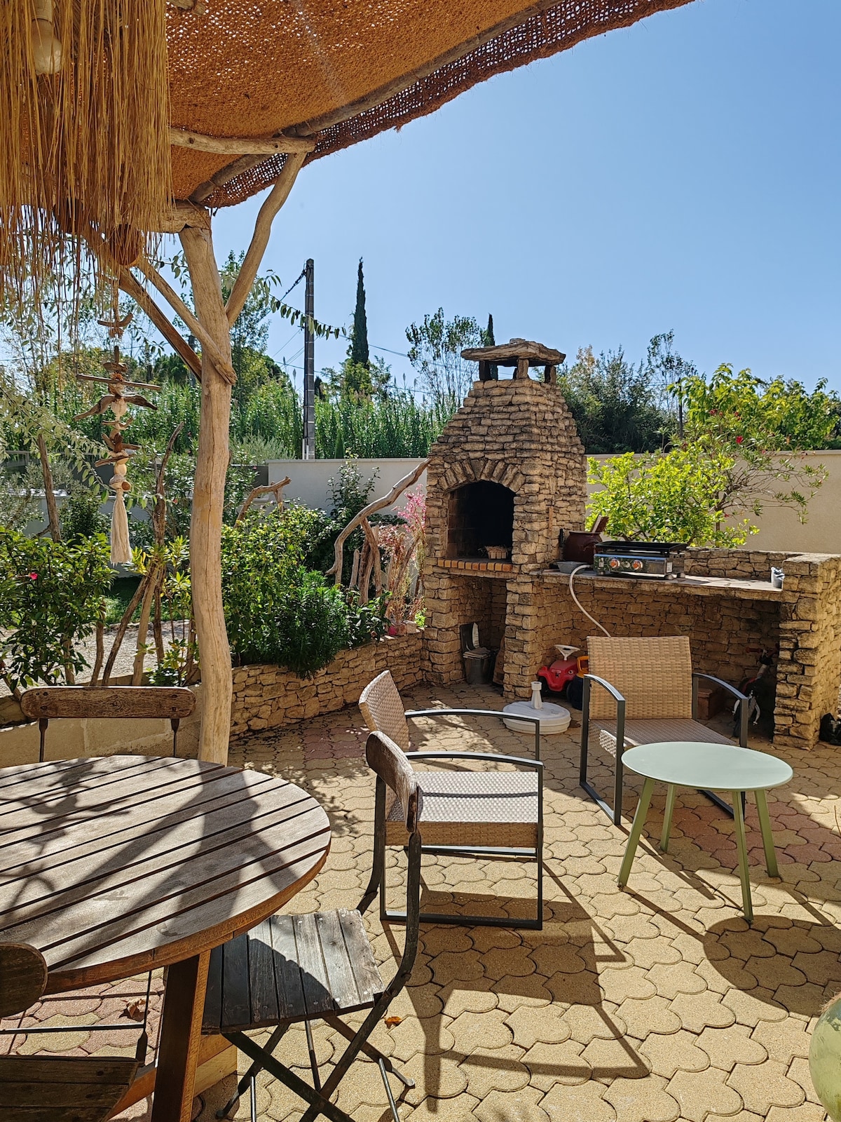 A private terrace is featured, showcasing a stone barbecue and a wooden table surrounded by four chairs. Lush greenery lines the background, complemented by a sunny blue sky. The space is designed for outdoor dining and relaxation.