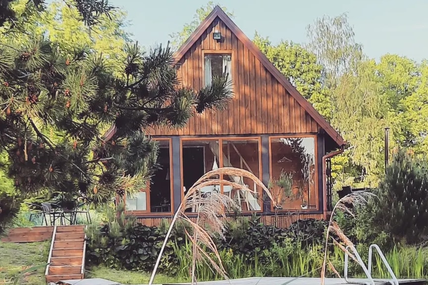 The two-story wooden cabin features a sloped roof and large windows framed by lush greenery. A wooden deck is visible, leading to the garden, which includes a variety of plants. A peaceful outdoor ambiance is created by the natural surroundings.