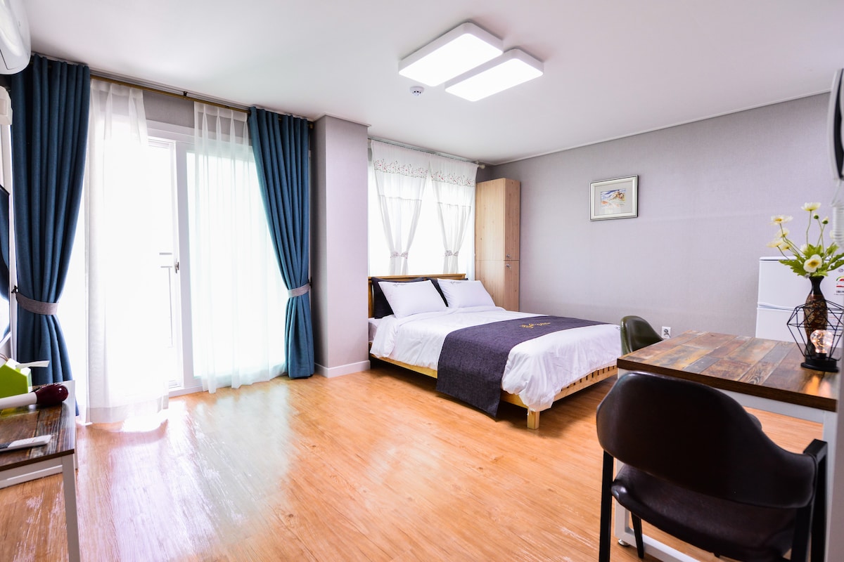 A bright room is featured with natural light flooding in through large curtains. A spacious bed with neutral bedding is positioned beside a modern wooden table and chair. A closet stands against the wall, complementing the minimalist décor.