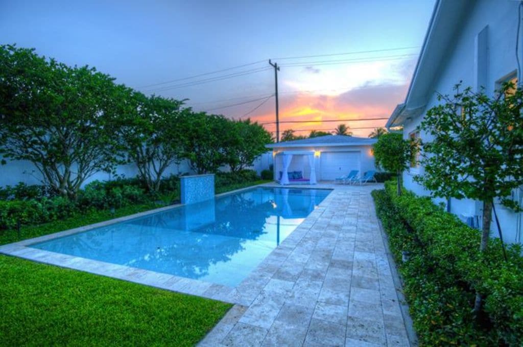 A serene backyard pool area is presented, featuring a well-maintained rectangular pool surrounded by lush greenery. The stone patio leads to a soothing sunset in the background, reflecting warm hues on the water's surface, creating a peaceful outdoor setting.