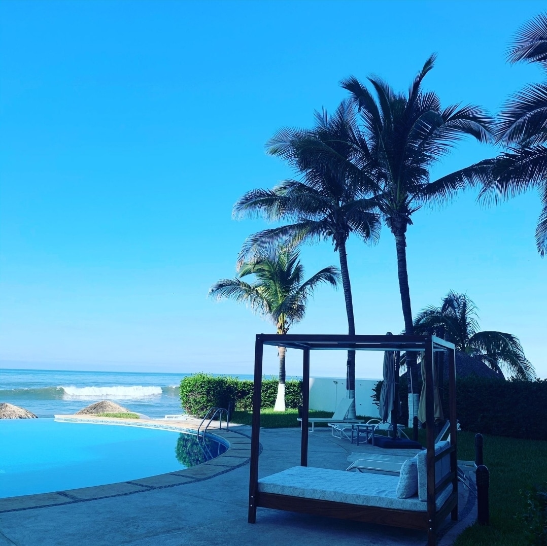 A view of a tranquil pool area surrounded by palm trees, with the ocean visible in the background. A shaded seating area features a cushioned bed, while loungers are positioned nearby. The clear blue sky adds to the serene environment, inviting relaxation.
