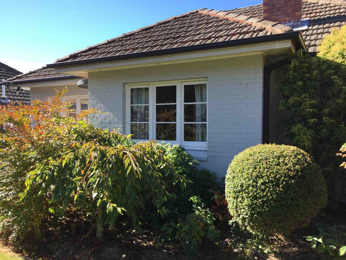 A charming exterior view of the house features a well-maintained garden with vibrant greenery. A large window framed by soft curtains allows natural light to illuminate the interior. The brick façade complements the surrounding foliage, enhancing the tranquil outdoor ambience.