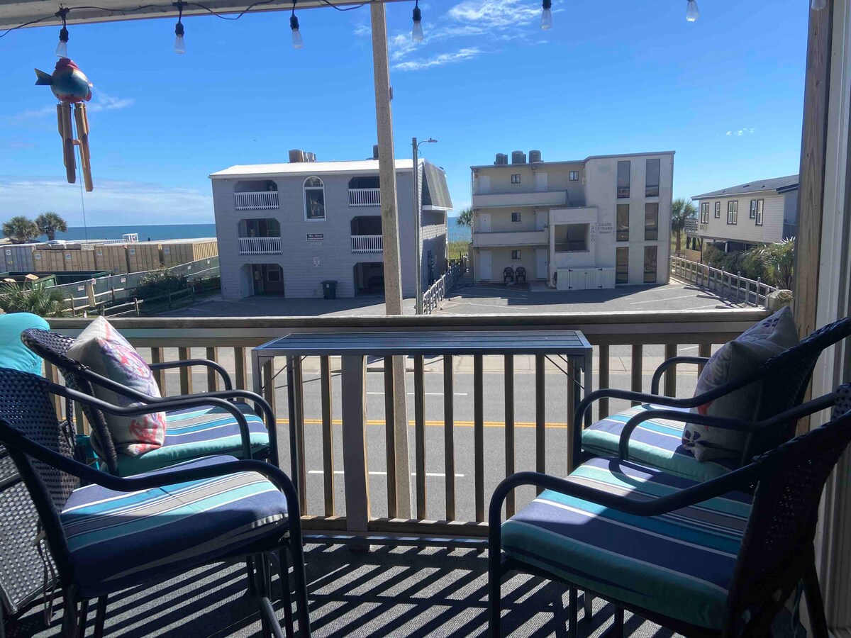 A private balcony view is showcased, featuring two chairs with vibrant cushions. The scene includes a clear blue sky, with a glimpse of the ocean visible in the distance. Nearby buildings and a street are visible, enhancing the coastal atmosphere.