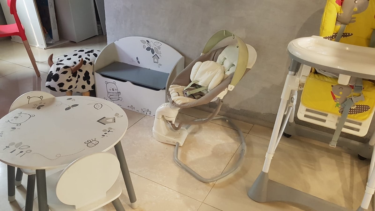 A child-friendly space includes a small table with two chairs, a cozy baby rocker, and a high chair. The items feature playful designs, while the surroundings have a neutral-toned wall and tiled flooring, creating a welcoming environment for families.
