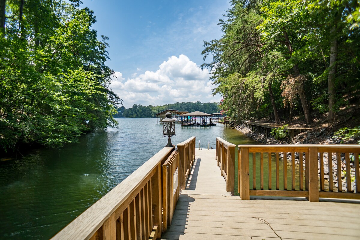 A wooden dock extends out over calm waters, surrounded by lush greenery and trees. The serene lake reflects a bright blue sky with fluffy clouds. A second dock can be seen in the distance, adding to the picturesque lakeside setting.