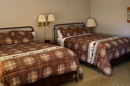 A room is furnished with two spacious queen beds, each layered with warm plaid bedding. Bedside lamps provide soft lighting. A subtle pattern adorns the carpet, while neutral-colored walls create a calming environment.