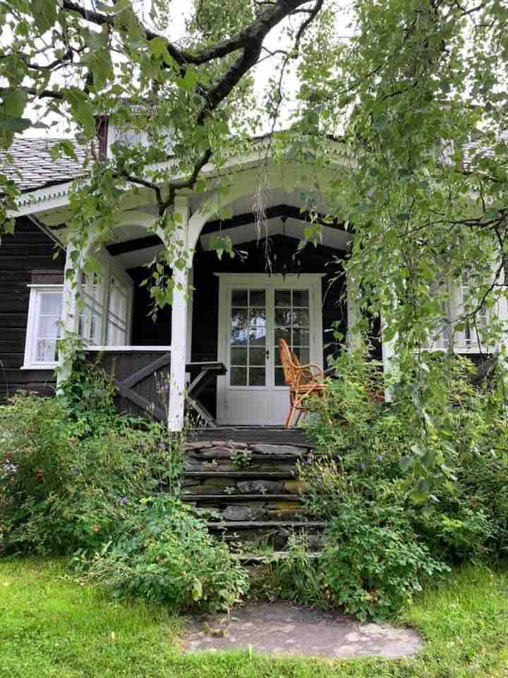 A welcoming entrance to a cabin is framed by lush greenery and a large tree overhead. Steps lead to a wooden porch, featuring a chair positioned beside glass-paneled doors. The surrounding garden adds a natural touch to the rustic charm of the setting.