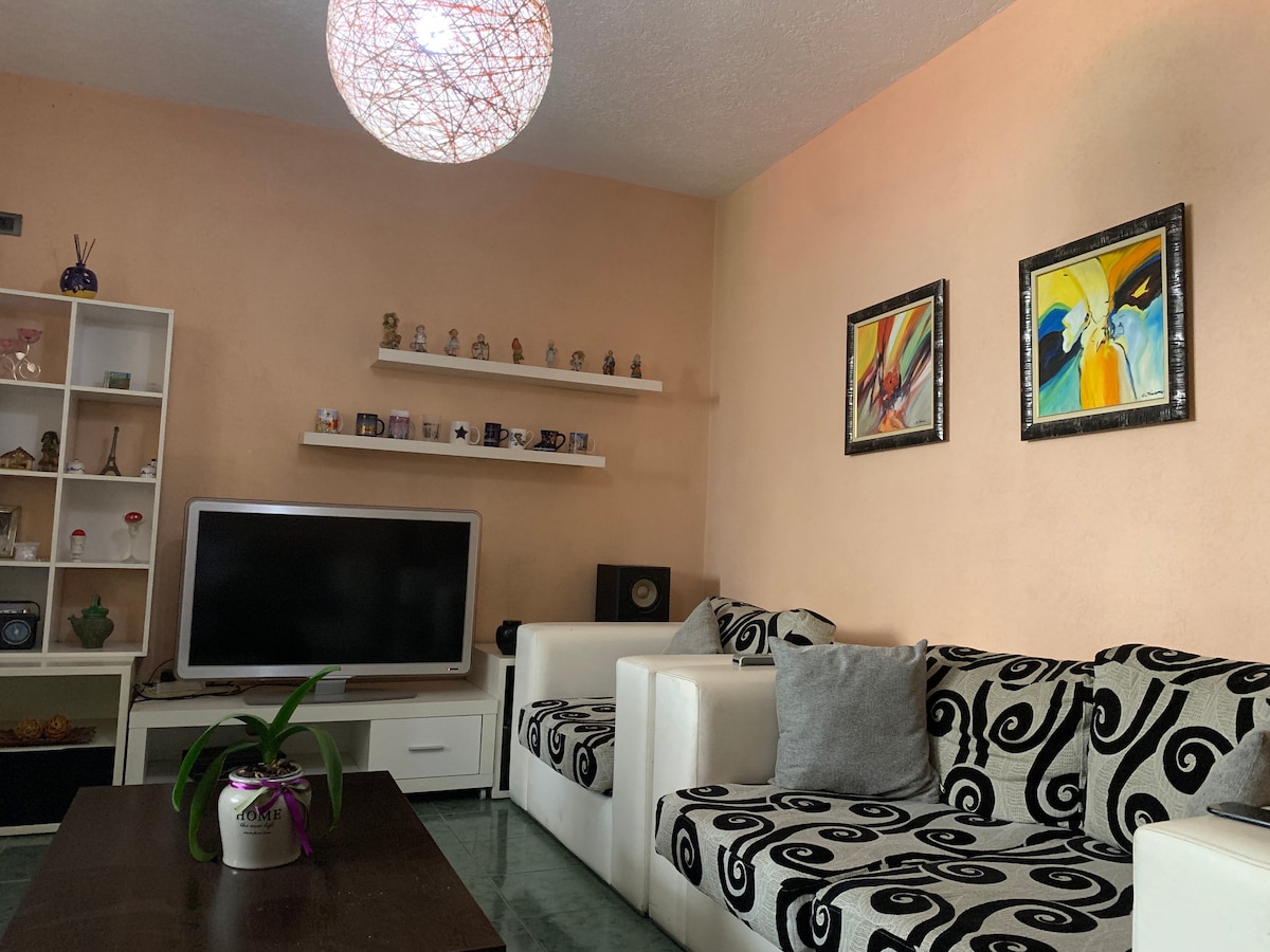 A cozy living area features contrasting black and white patterned sofas positioned around a wooden coffee table. A flat-screen television is mounted on the wall below a shelf displaying various decorative items. Artwork adorns the walls, while a unique light fixture hangs from the ceiling.